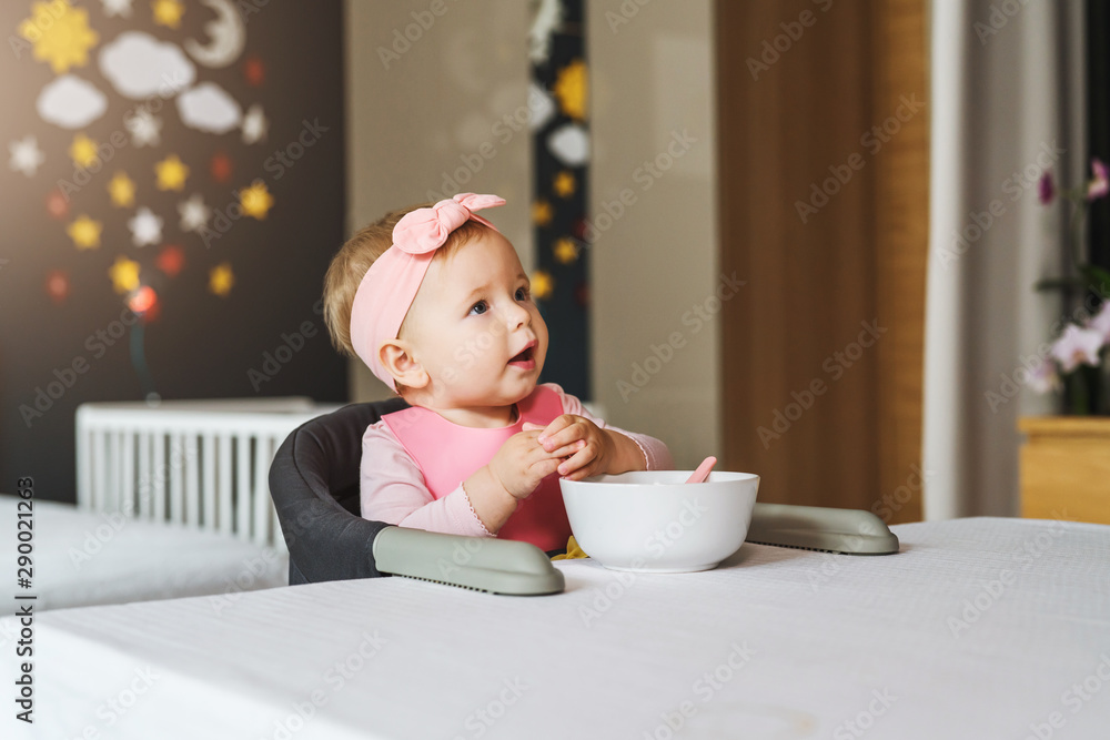 Nine-month-old smiling baby girl sits at white table in highchair and eats herself with spoon from bowl. Blurred background. Nine-month-old smiling baby girl sits at white table in highchair and eats herself with spoon from bowl. Blurred background.