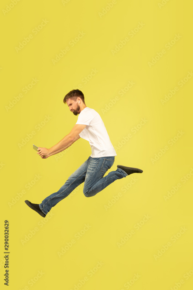 Full length portrait of happy jumping man isolated on yellow background. Caucasian male model in casual clothes. Freedom of choices, inspiration, human emotions concept. Takes video for vlog or