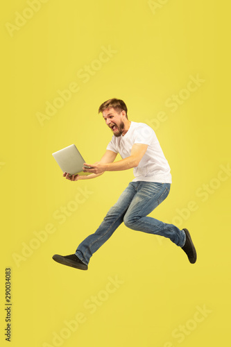 Full length portrait of happy jumping man isolated on yellow background. Caucasian male model in casual clothes. Freedom of choices, inspiration, human emotions concept. Using tablet in flight. © master1305
