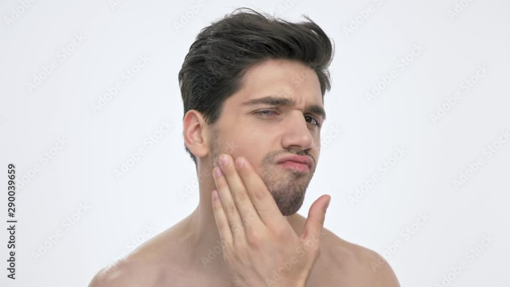 Close up view of serious young brunette man with naked torso looking at his bristle and thinking about shaving while looking at the camera over white background isolated Close up view of serious young brunette man with naked torso looking at his bristle and thinking about shaving while looking at the camera over white background isolated