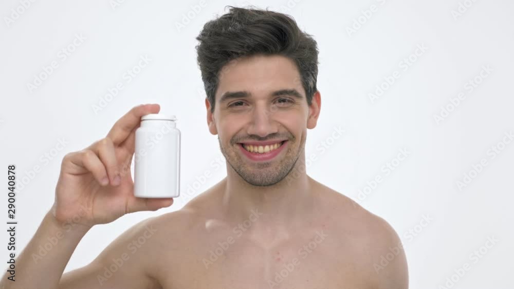Close up view of smiling young brunette man with naked torso showing a bottle with pills or vitamins while looking at the camera over white background isolated Close up view of smiling young brunette man with naked torso showing a bottle with pills or vitamins while looking at the camera over white background isolated