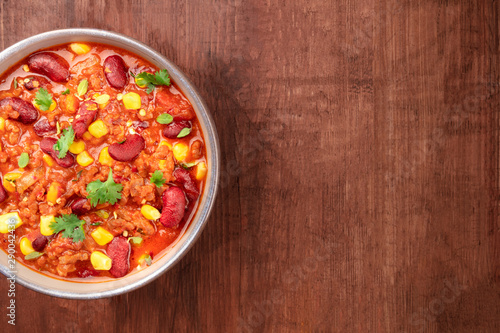 Chili con carne, a Mexican stew with red beans, cilantro leaves, ground beef, and chili peppers, close-up overhead shot on a dark rustic wooden background with copy space