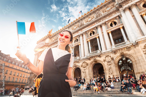 happy Asian girl traveler with french flag near the main Facade of the Opera Garnier in the historic building of the Academy of music of Paris. Blurred unrecognizable people at the background photo