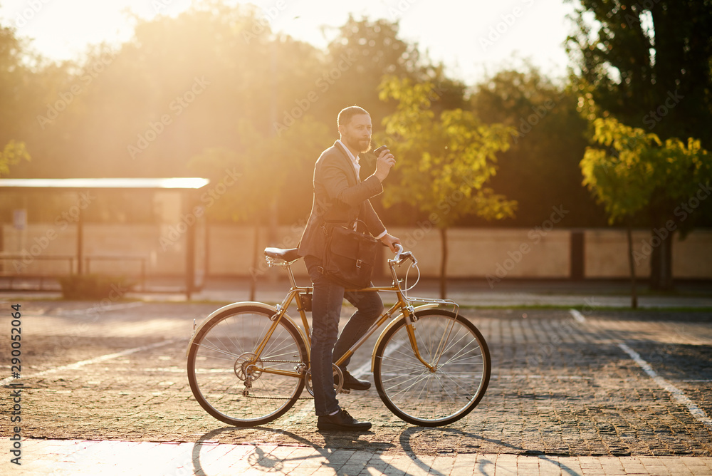 Obraz premium Portrait of handsome young red bearded man in casual clothes using phone, drink coffee and looking away and smiling while leaning on his bike, standing outdoors. Office and business center background