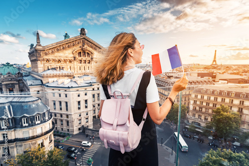 Happy asian woman with french flag and backpack standing on a roof top and enjoys great view over Parisian skyline at sunset. Travel and education in France © EdNurg
