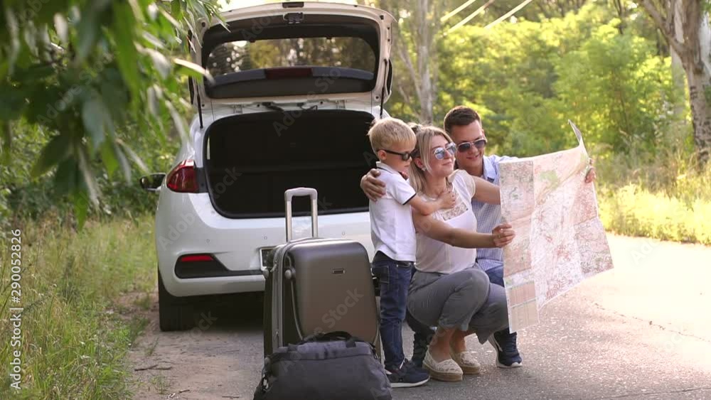 A young active family of travelers in sunglasses view a map near their car on an empty road in the woods. Summer trip by car. Husband, wife and child traveling by car. A young active family of travelers in sunglasses view a map near their car on an empty road in the woods. Summer trip by car. Husband, wife and child traveling by car.
