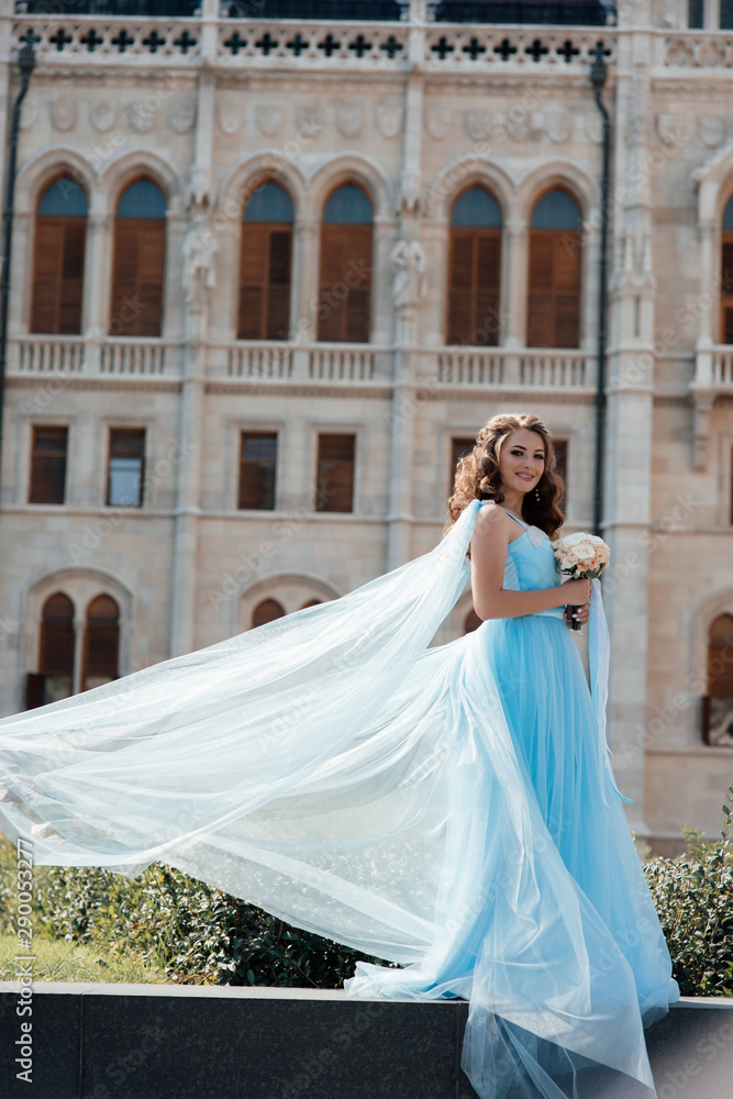 Fototapeta premium Elegant romantic woman in blue sexy dress with flowers standing near the buiding in Prague. Gorgeous girl enjoying. Dressed in blue fluttering dress. Pretty woman with makeup and hairstyle