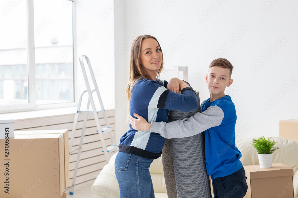 Fototapeta premium Relocation, housewarming and real estate concept - Smiling single mother and a charming son are holding a folded carpet in a new living room intending to spread it in a new apartment