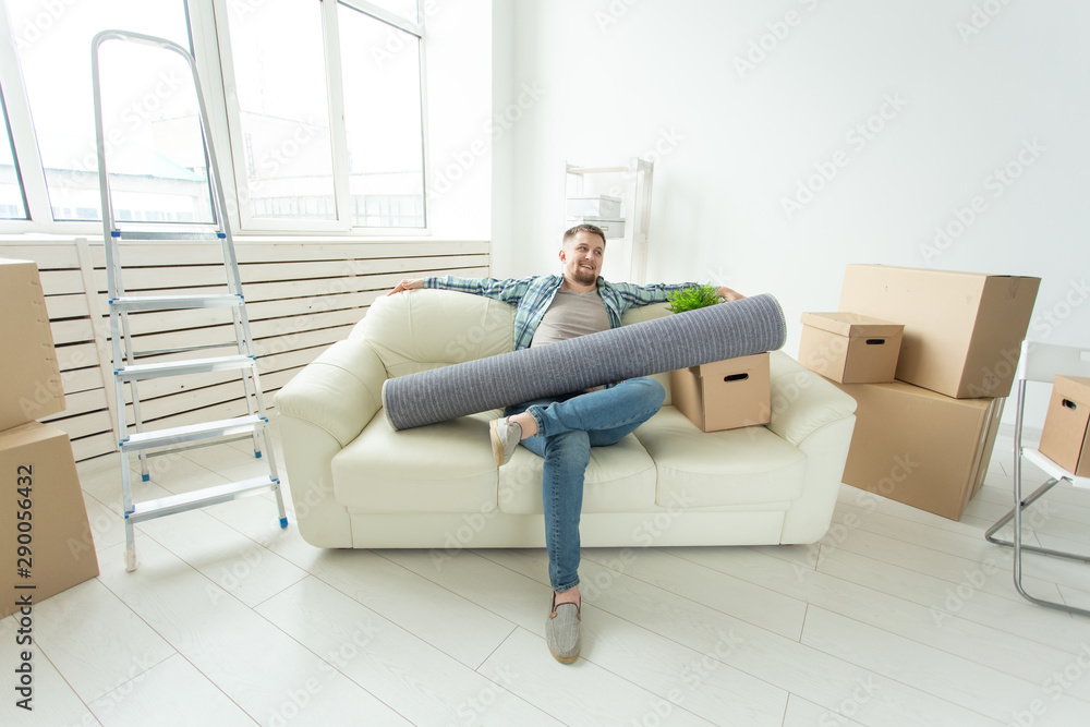 Positive young man sitting on the sofa with his things in the new living room after the move. Concept of housing affordability for young people. Positive young man sitting on the sofa with his things in the new living room after the move. Concept of housing affordability for young people.