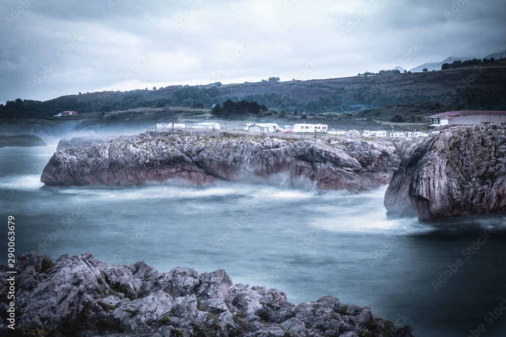 Agua con efecto seda con olas al lado de unos acantilados y montañas de fondo, con un parking para furgonetas camping encima Agua con efecto seda con olas al lado de unos acantilados y montañas de fondo, con un parking para furgonetas camping encima