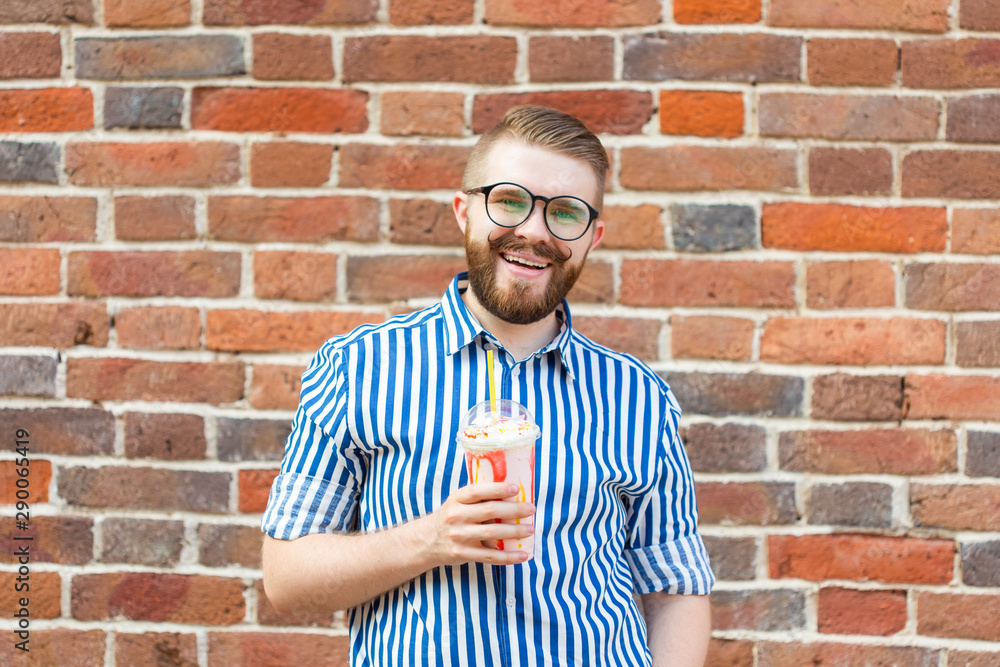 Blurred positive young hipster man with beard and mustache in glasses holding a fruit milkshake in his hands posing on the street near a brick wall. Concept of summer walks. Blurred positive young hipster man with beard and mustache in glasses holding a fruit milkshake in his hands posing on the street near a brick wall. Concept of summer walks.