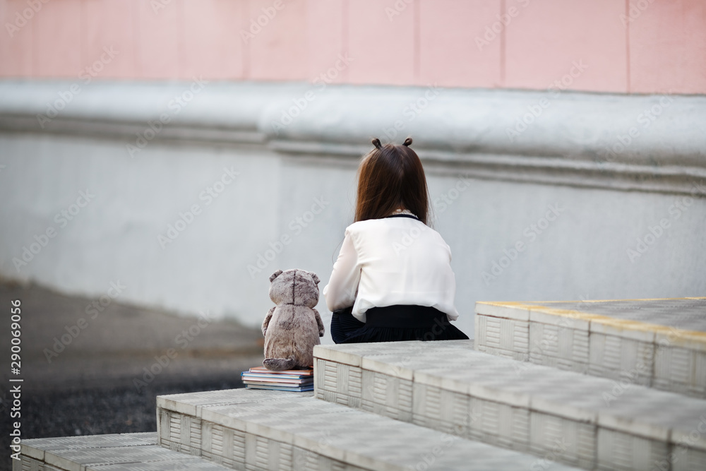 Φωτογραφία Sad girl with a plush toy sits on the steps of the school stairs with her back to the camera