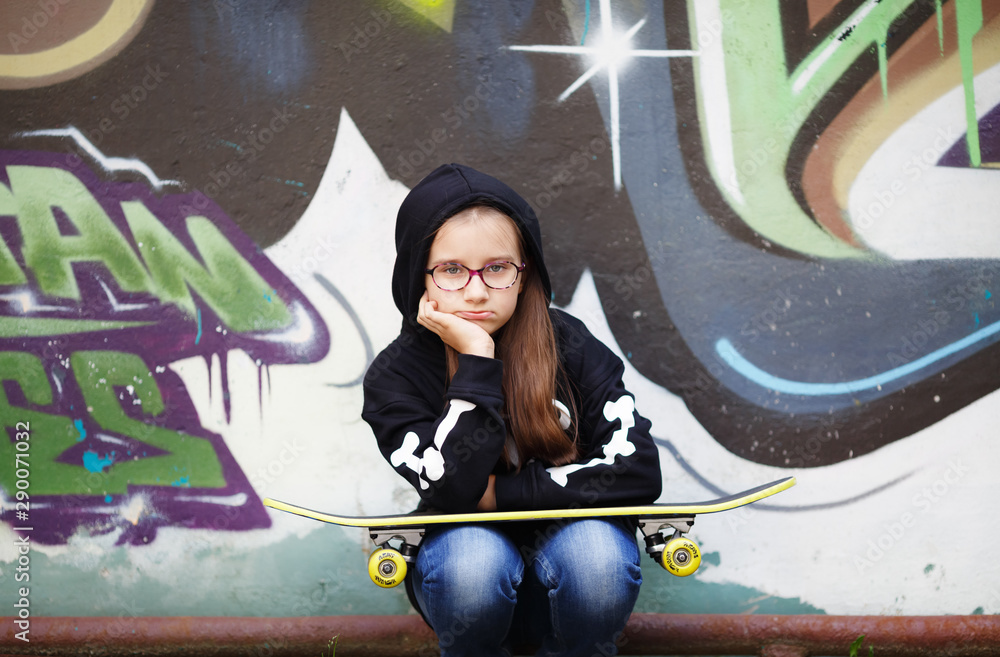 Child girl sitting with skateboard on her knees against wall background. Selective focus.