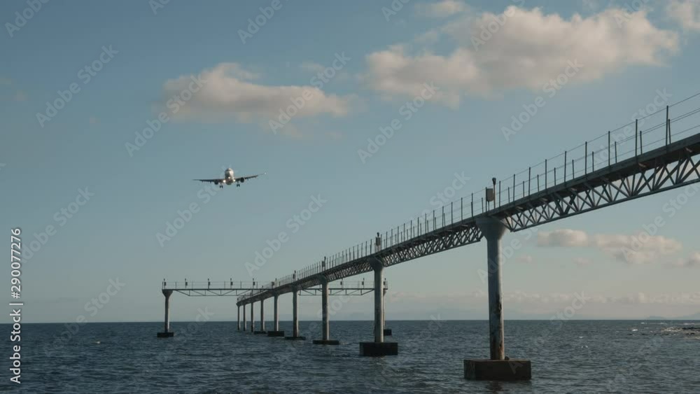 A plane is flying in a bright blue sky low over the calm sea. It is flying over a large framed pipeline standing in the water A plane is flying in a bright blue sky low over the calm sea. It is flying over a large framed pipeline standing in the water