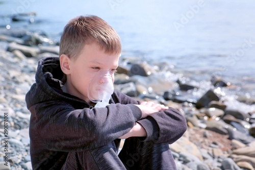 Portrait sick 8y child boy uses nebulizer sits on seaside. Inhaling inhaler mask. Fibrosis cystic copd and treatment. Asthma pulmonary respiratory breath problem cure. Painkiller sedative gas oxygen.