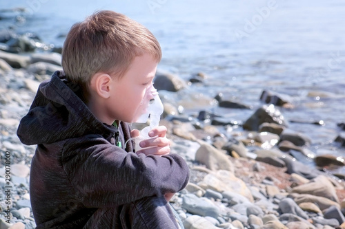 Sick child boy uses nebulizer sits on seaside, side view. Inhaling inhaler mask. Fibrosis cystic copd and treatment. Asthma pulmonary respiratory breath problem cure. Painkiller sedative gas oxygen.