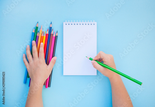 Child's hands holding colorful pencils and a notebook on blue background with copyspace. Flat lay style. Back to school concept.