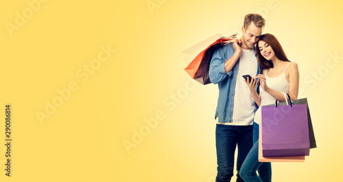 Picture of happy smiling lovely couple with smartphone and shopping bags, against yellow color background