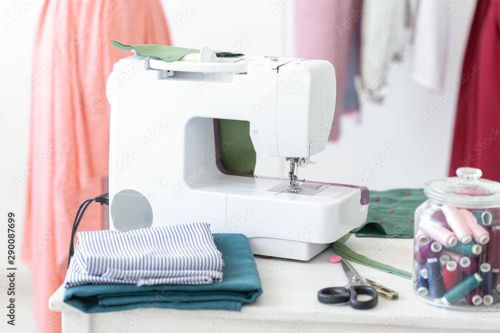 Designer's desk sewing machine thread fabric scissors against the background of finished stitched products. Concept of tailoring to order.