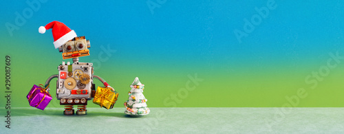 Christmas New Year robotic greeting card mockup. Funny robot Santa Claus holds gift boxes. Toy pine tree on blue green wall gray floor background. copy space for congratulations holidays text.
