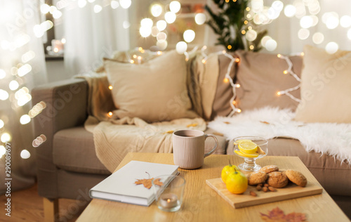 hygge and cozy home concept - cup of tea with lemon, book, autumn leaves and oatmeal cookies on wooden table in living room