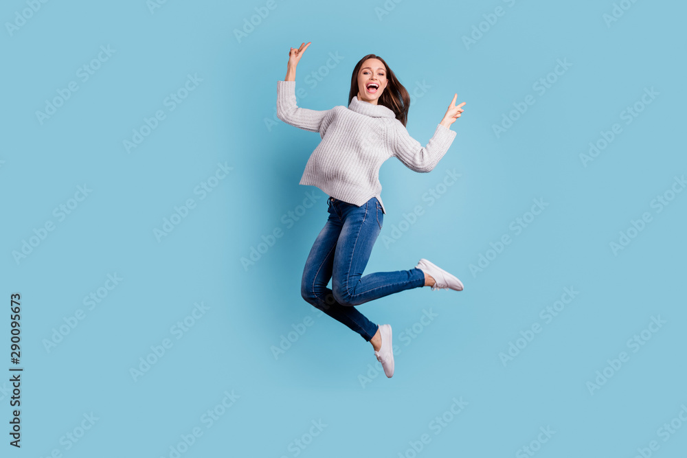 Full length photo of content candid girl jump have free time make v-signs feel crazy wear denim jeans white pullover white sneakers isolated over blue color background Full length photo of content candid girl jump have free time make v-signs feel crazy wear denim jeans white pullover white sneakers isolated over blue color background