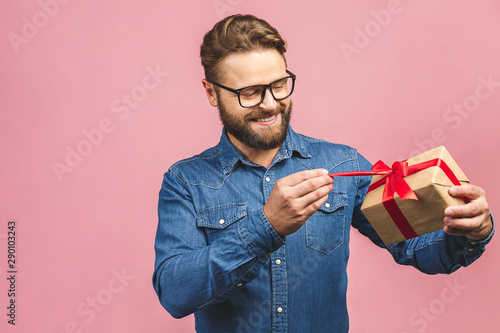 Happy holiday, my congredulations! Portrait of an attractive casual man giving present box and looking at camera isolated over pink background.
