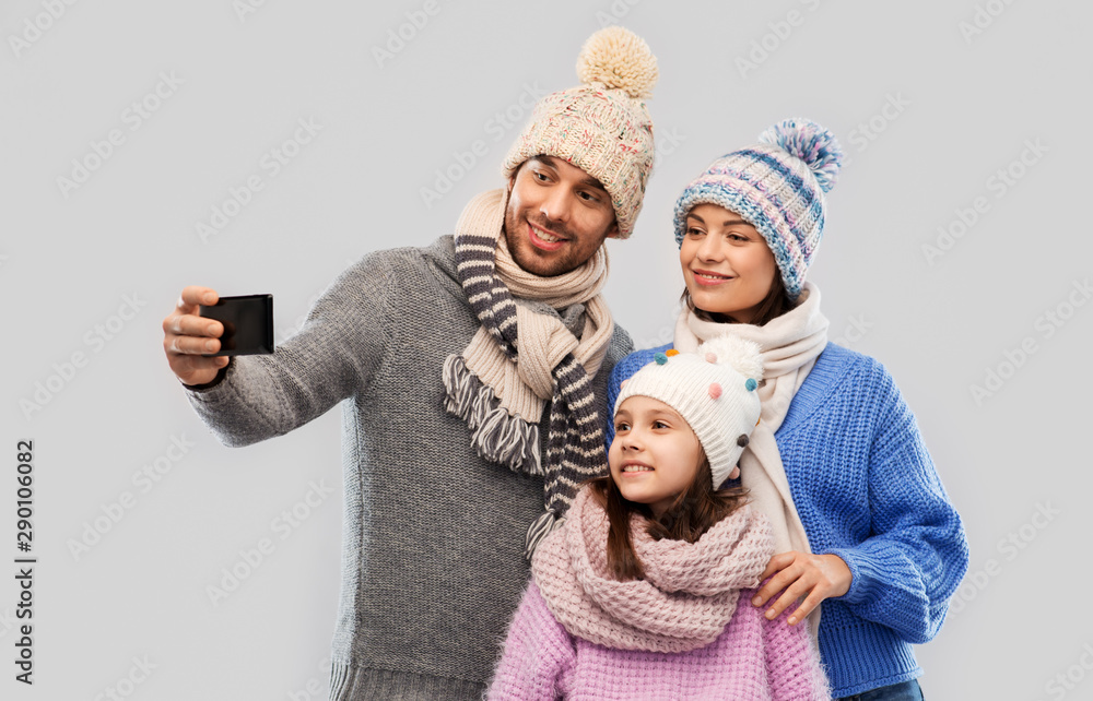 family, christmas and technology concept - happy mother, father and little daughter in winter clothes taking selfie by smartphone over grey background family, christmas and technology concept - happy mother, father and little daughter in winter clothes taking selfie by smartphone over grey background