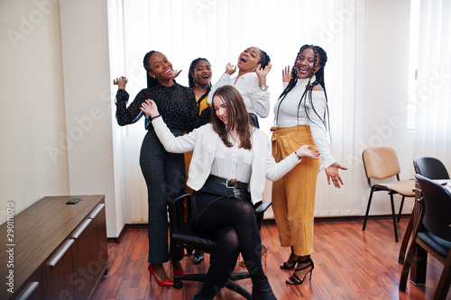 Five multiracial business womans standing at office and roll woman on chair. Diverse group of female employees in formal wear having fun.