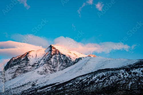 Pink light of sunrise hitting the top of a snow covered mountain with clouds and blue sky