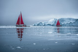 Beautiful red sailboat in the arctic next to a massive iceberg showing the scale. Cruising among floating icebergs in Disko Bay glacier during midnight sun season of polar summer Ilulissat, Disko Bay