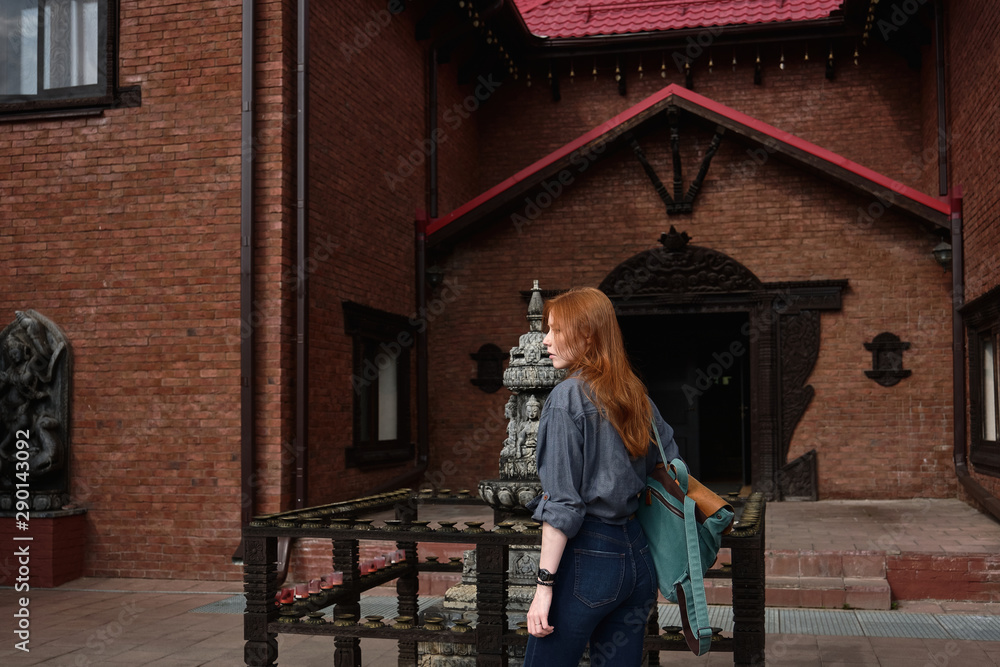 A red-haired girl in a denim shirt with a backpack stands half a turn in the courtyard of an Asian temple.
