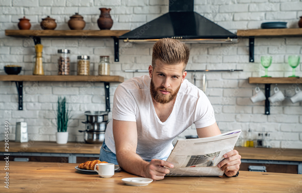 Starting day from good news. Handsome young man reading newspaper seriously while drinking coffee and having breakfast in the kitchen, and looning to the camera Starting day from good news. Handsome young man reading newspaper seriously while drinking coffee and having breakfast in the kitchen, and looning to the camera