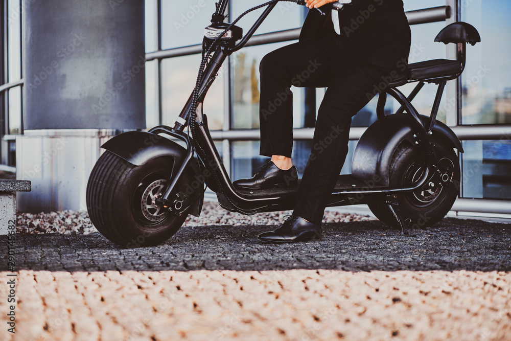 Fototapeta premium Elegant businessman in black suit is sitting on his electrical scooter near his office.