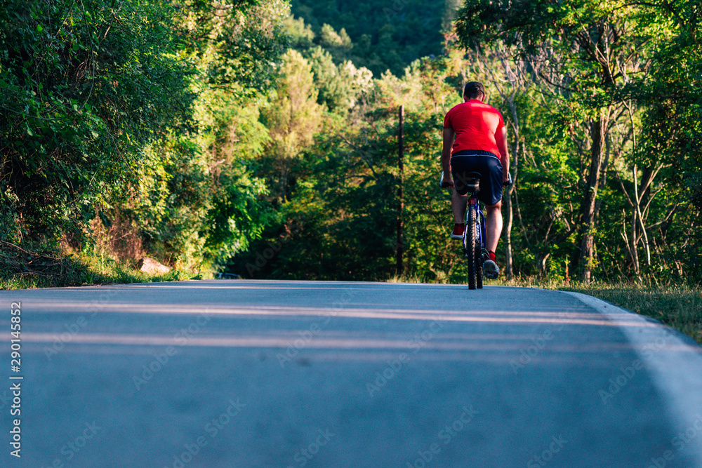 Fototapeta premium Fit cyclist rides his bicycle (bike) on an empty road in nature wearing a baseball hat and red t-shirt.
