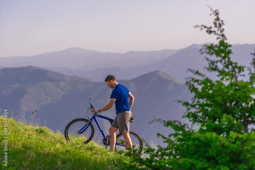 Tired cyclist is wiping his sweat off his face while pushing his bicycle uphill on a dirt road in a mountain.