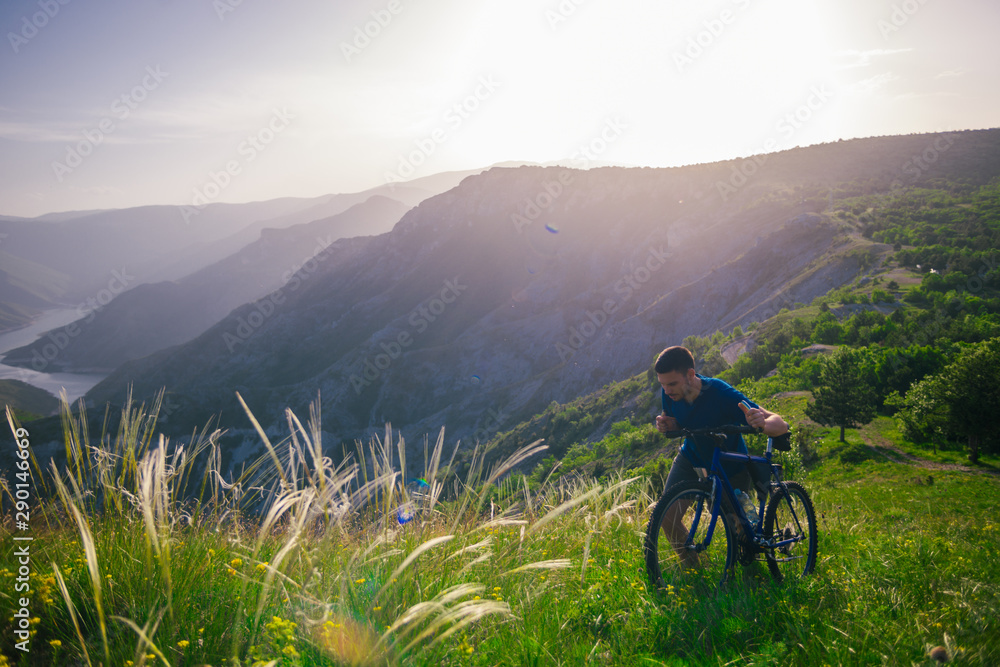 Obraz premium Perspective of a fit mountain biker pushing his bike uphill with amazing view on a forest, river and mountains in the background. Amazing green nature at sunset.