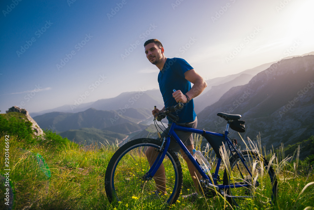 Fototapeta premium Perspective of a fit mountain biker pushing his bike uphill with amazing view on a forest, river and mountains in the background. Amazing green nature at sunset.