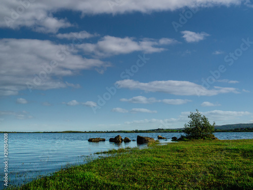 Blue surface of lake Corrib and cloudy sky, Grass covers shore. Warm sunny day. © mark_gusev