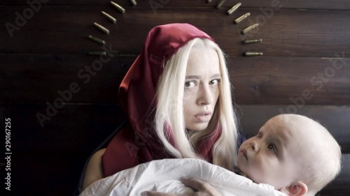 Woman with the covered head, mother with a baby in her arms standing against wooden wall with an arc of bullets, religion and war concept. Footage. The image of the virgin Mary and a Jesus.