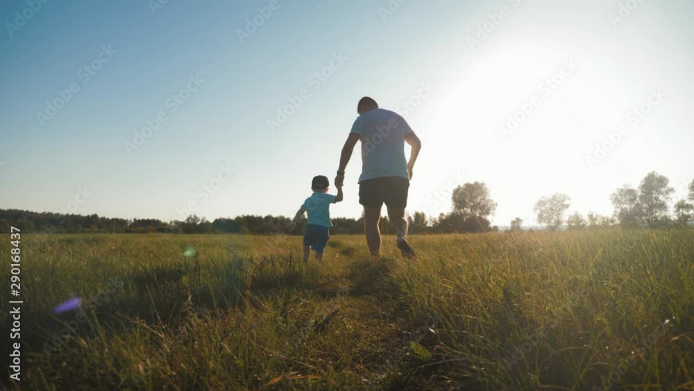 Happy male and boy running on sunlit meadow. Dad and his kid walking on trail among field. Concept of a happy family having fun during the summer activity. Happy male and boy running on sunlit meadow. Dad and his kid walking on trail among field. Concept of a happy family having fun during the summer activity.