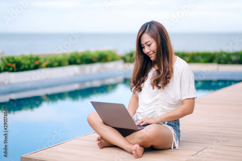 A beautiful asian woman using and typing on laptop computer while sitting by swimming pool