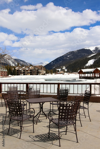 Colorado nature background, ski resort vacation concept. Scenic view with outdoor furniture on a terrace in from of covered by melting ice pond, snowy mountains and accommodations for vacation rental.