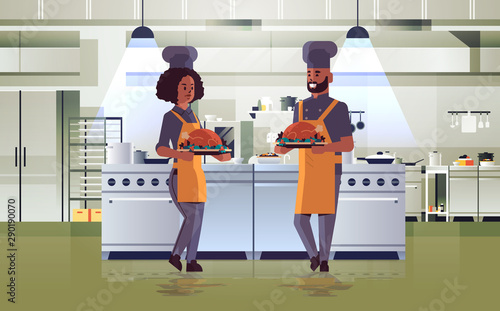 professional chefs couple holding trays with roasted chicken man woman in uniform carrying thanksgiving turkey cooking food concept modern restaurant kitchen interior full length horizontal
