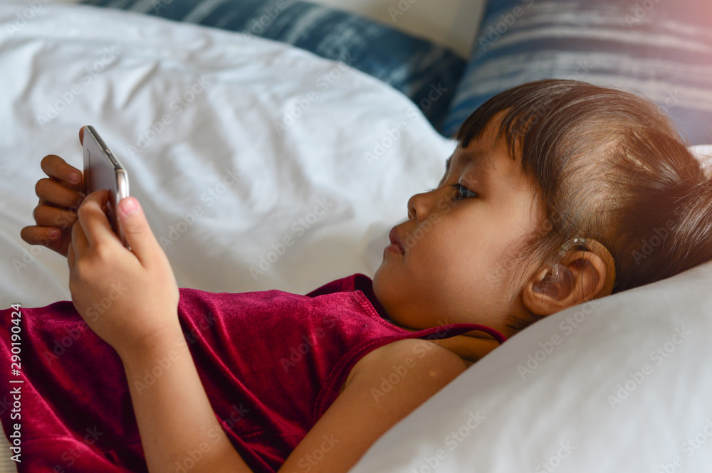 Asian children girl hearing impaired and put on hearing aids play smartphone on the bed. Asian children girl hearing impaired and put on hearing aids play smartphone on the bed.