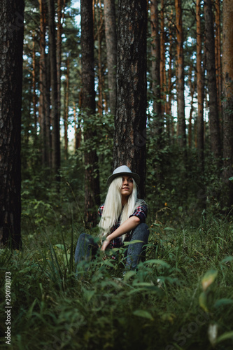 Young adult blonde female in a hat sitting on the ground in the woods, selective focus