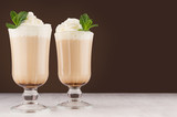 Refreshment coffee drink with fresh green mint, whipped cream, foam in two glass in luxury dark brown interior on white wood table, copy space.
