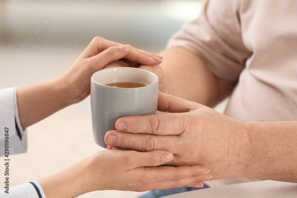 Fototapeta premium Nurse giving cup of tea to elderly man against blurred background, closeup. Assisting senior generation