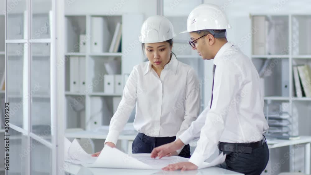 Medium shot of Asian female and Middle Eastern male engineers in business attire and white hardhats standing in office, studying technical drawings on large paper sheets and discussing details. Medium shot of Asian female and Middle Eastern male engineers in business attire and white hardhats standing in office, studying technical drawings on large paper sheets and discussing details.