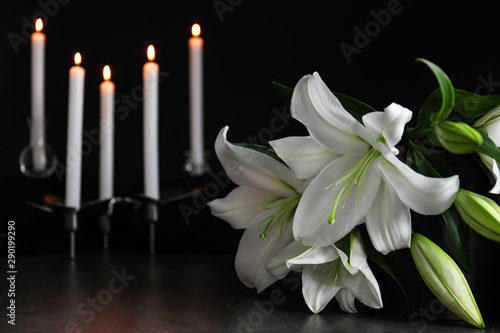 White lilies and blurred burning candles on table in darkness  closeup with space for text. Funeral symbol